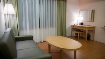 A hotel room with a green sofa, a wooden table, a desk with a chair, a lamp, and a large mirror on the wall.