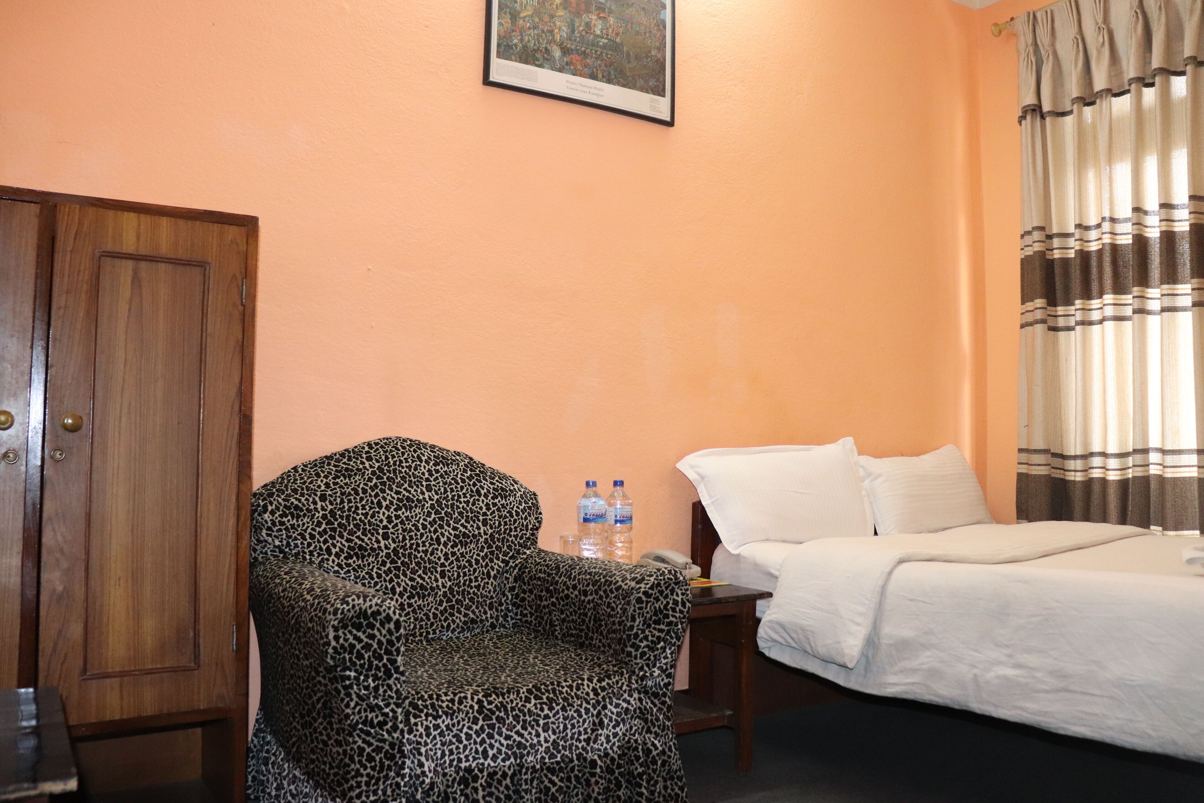Una habitación de hotel con una cama, un sillón estampado de leopardo, una puerta de madera, una mesita con botellas de agua y un cuadro enmarcado en la pared.