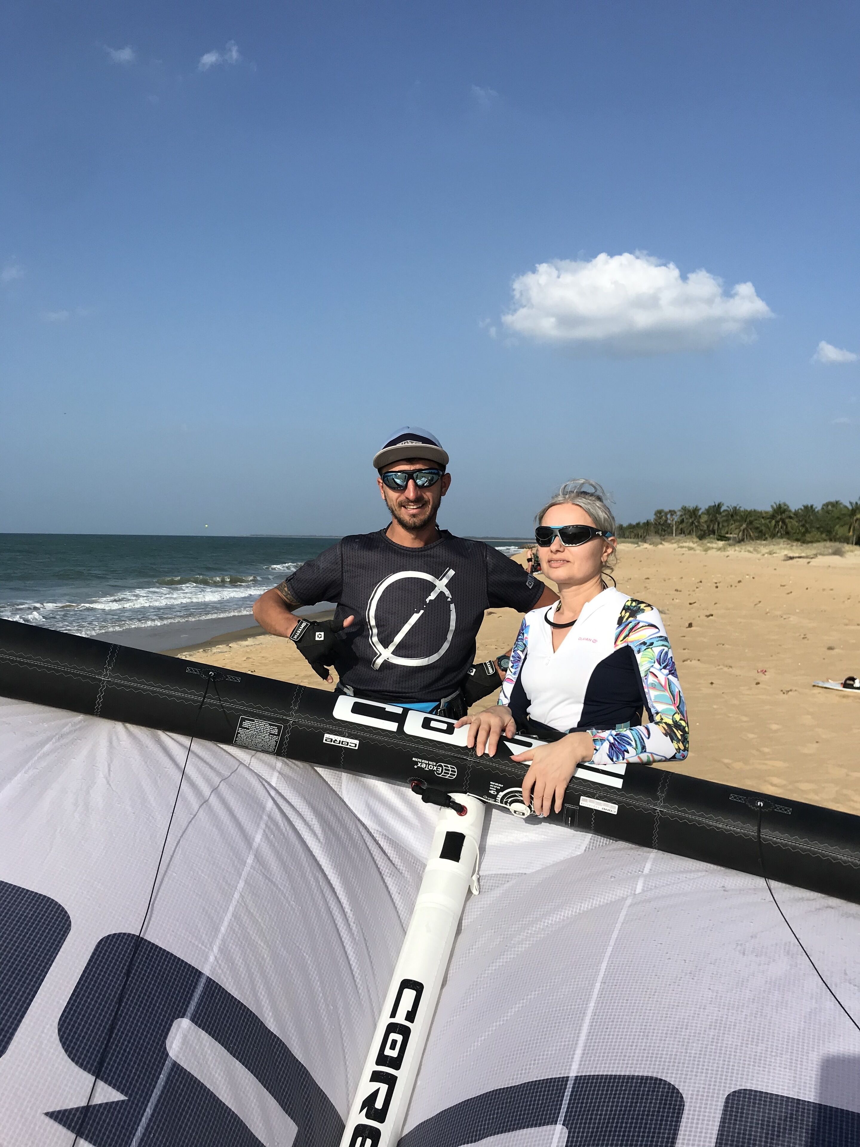 Two people standing by a large kiteboard on a beach with the ocean in the background.