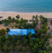 Vista aérea de uma praia com um prédio de telhado azul, rodeado por palmeiras.