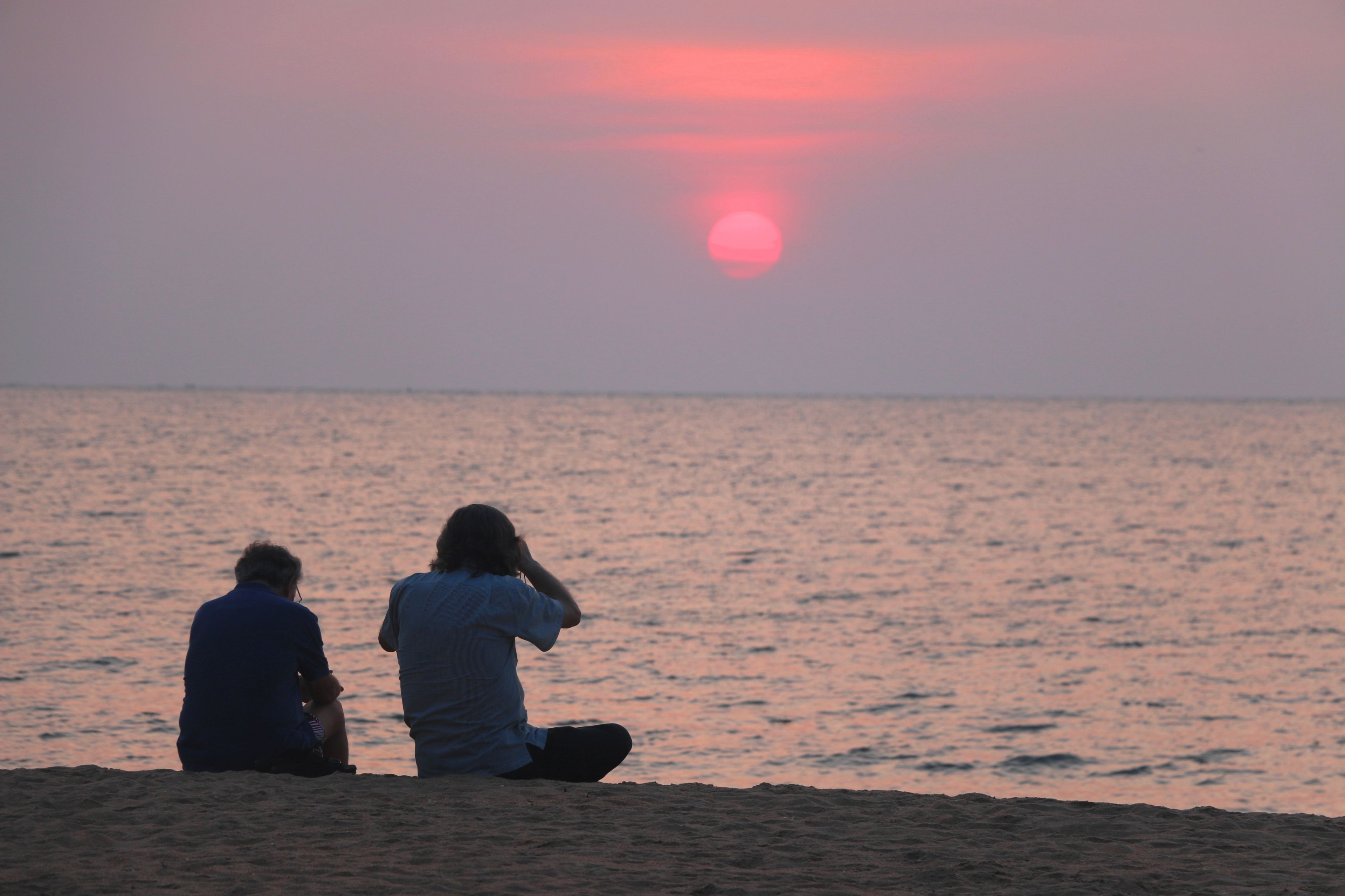 Zwei Personen sitzen am sandigen Strand und blicken zum Meer bei Sonnenuntergang.