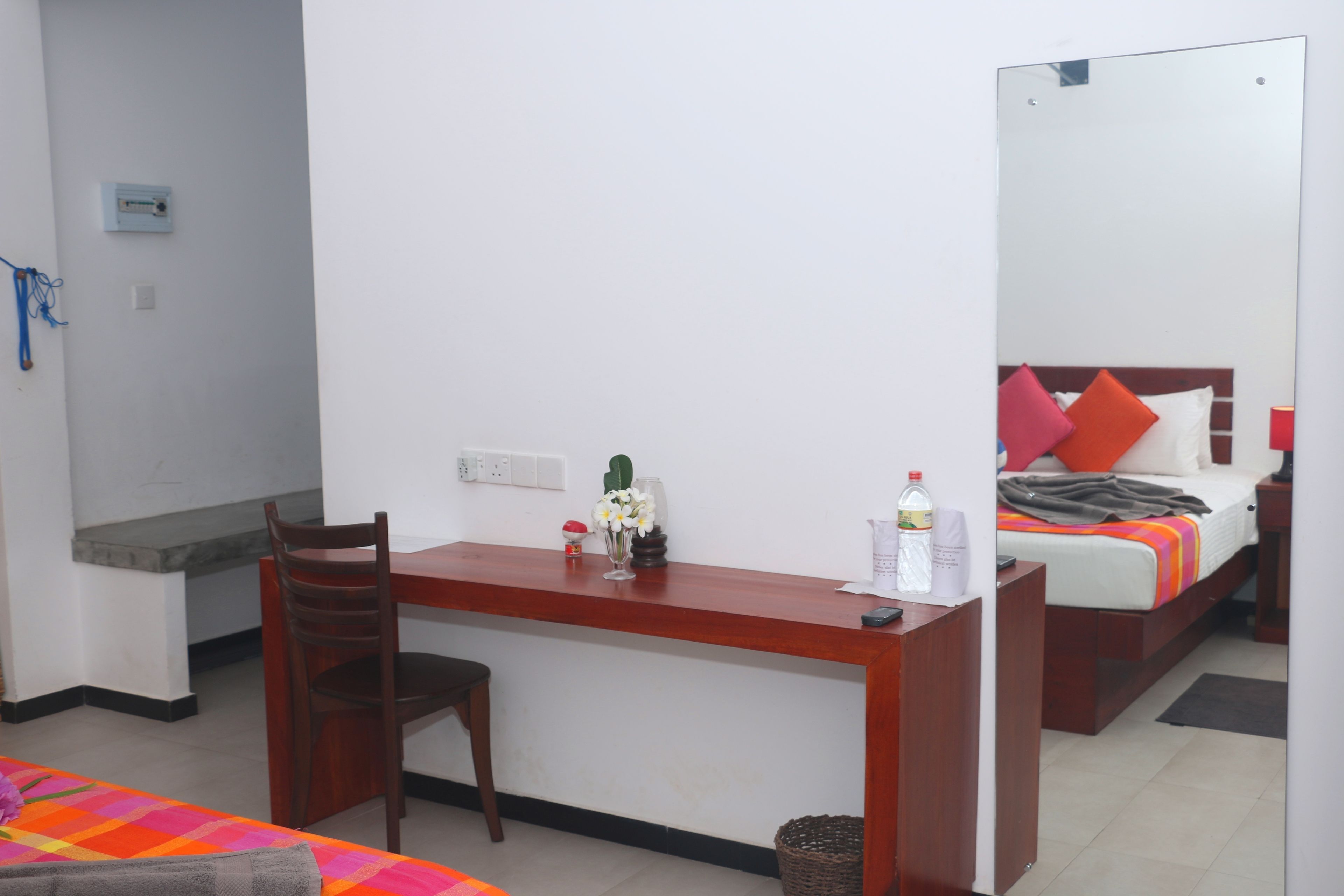 A hotel room with a wooden desk, a chair, a bed with colorful bedding, a bedside table, and a mirror.