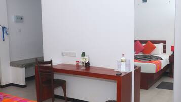 A hotel room with a wooden desk, a chair, a bed with colorful bedding, a bedside table, and a mirror.