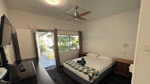 A hotel room with a bed, a desk with a computer, a ceiling fan, and a window with a view of greenery. - Cairns City Motel (Cairns)