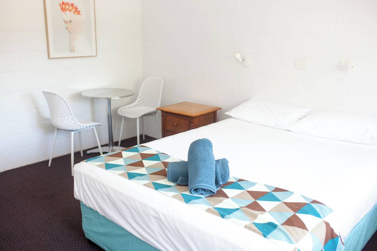 Una habitación de hotel con cama, dos sillas, una mesita y una lámpara fijada a la pared.