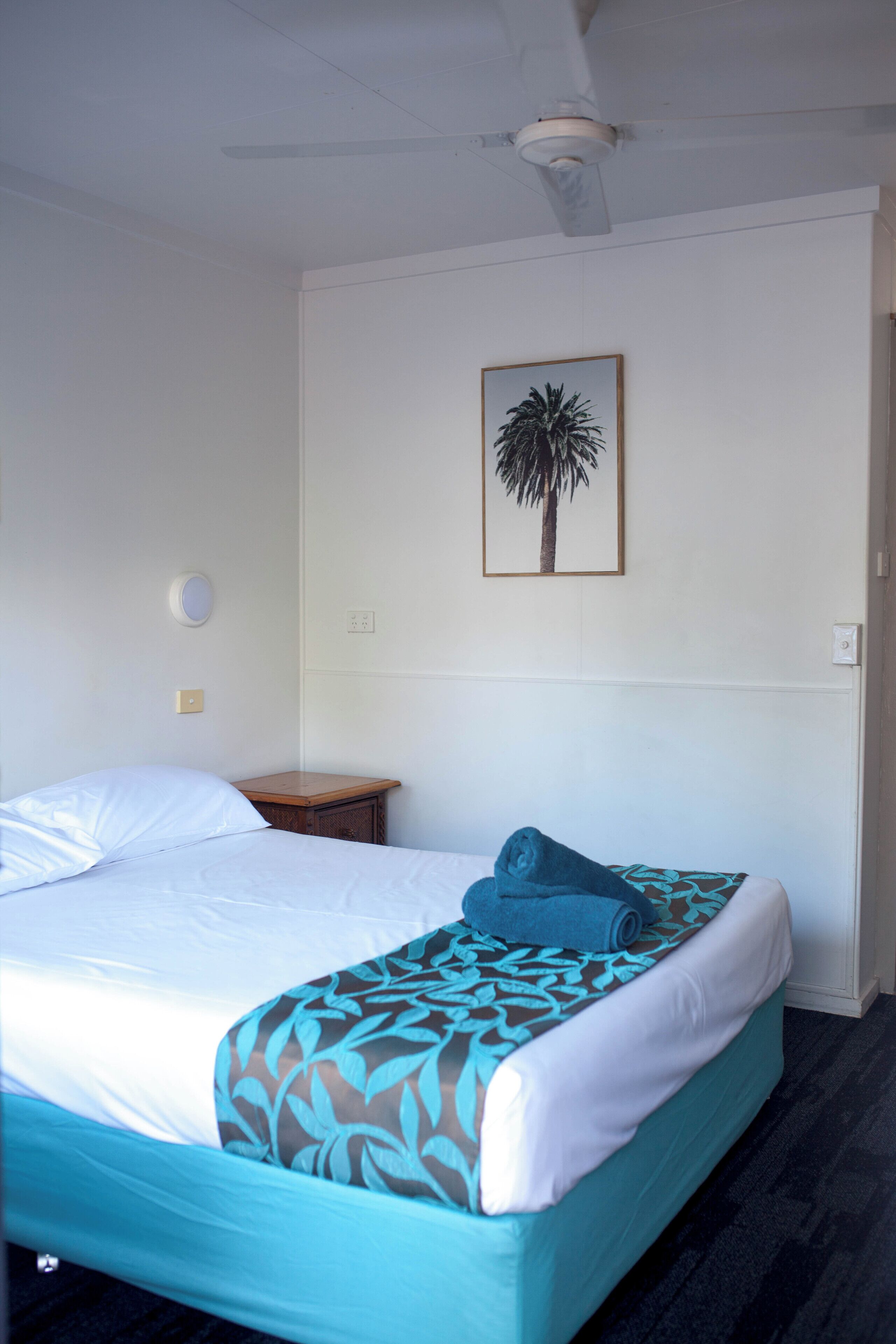 Hotelkamer met een bed, nachtkastje, een wandlamp en een lijst met een afbeelding van een palmboom.