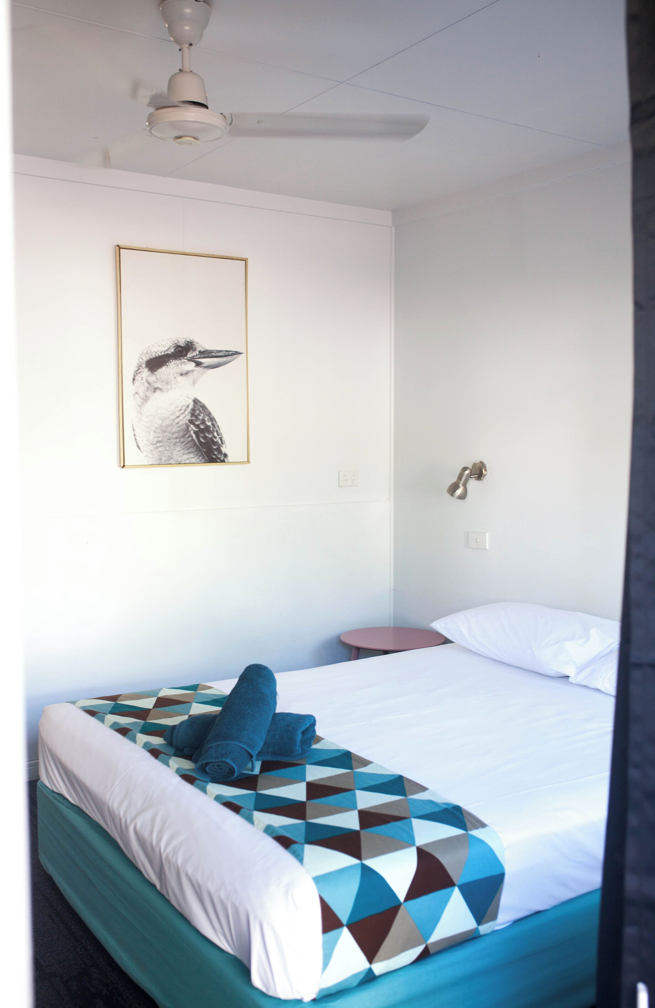 A hotel room with a bed, a small bedside table, a wall-mounted light, a ceiling fan, and a framed picture on the wall.
