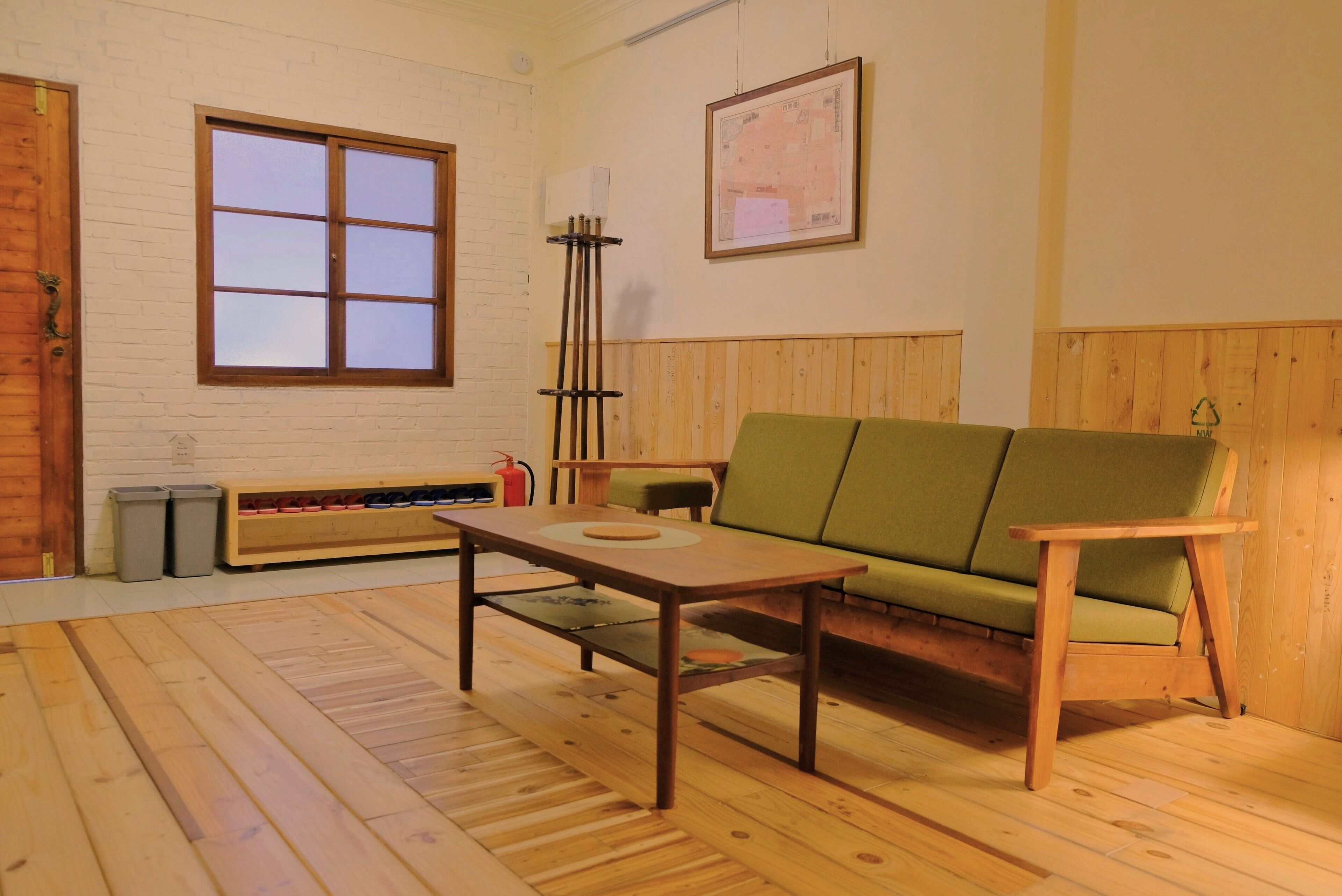 Ein Zimmer mit Holzboden, einem grünen Sofa, einem Holztisch, einem Fenster, einer an der Wand hängenden Karte und einem Mülleimer.