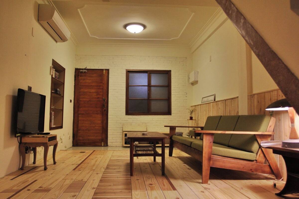 A living room with a wooden floor, a green sofa, a wooden coffee table, a TV on a stand, and a door with frosted glass panels.