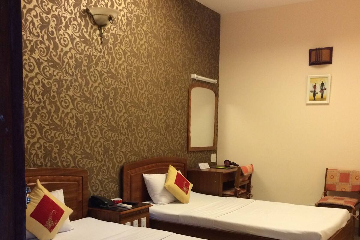 Ein Hotelzimmer mit zwei Einzelbetten, einem Nachttisch mit Lampe, einem Sessel und einem kleinen Schreibtisch mit Stuhl.