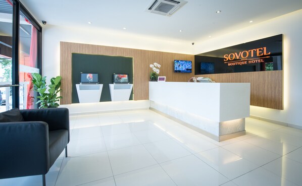 A modern hotel reception area with a reception desk, two monitors, a black sofa, and a potted plant.