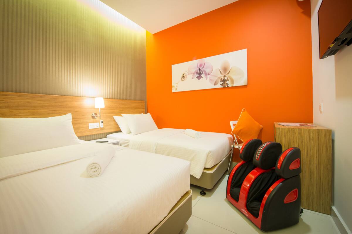 Ein Hotelzimmer mit zwei Betten, einem Massagesessel, einem Holzkopfende, einer orangefarbenen Akzentwand und einem Gemälde.