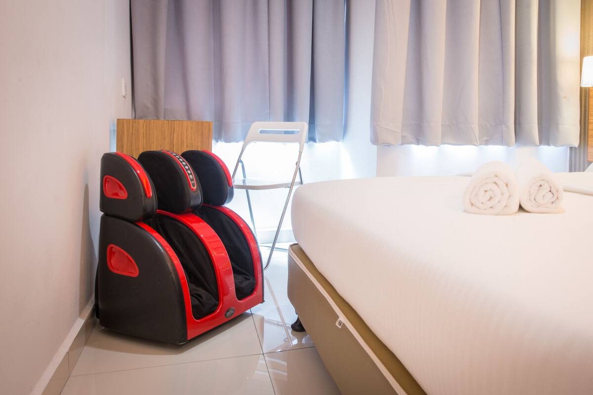 Ein Hotelzimmer mit Bett, zwei aufgerollten Handtüchern, einem Massagesessel und einem Fenster mit Vorhängen.