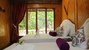 A room with two beds, wooden walls, a large window with a view of greenery, and a wooden door.