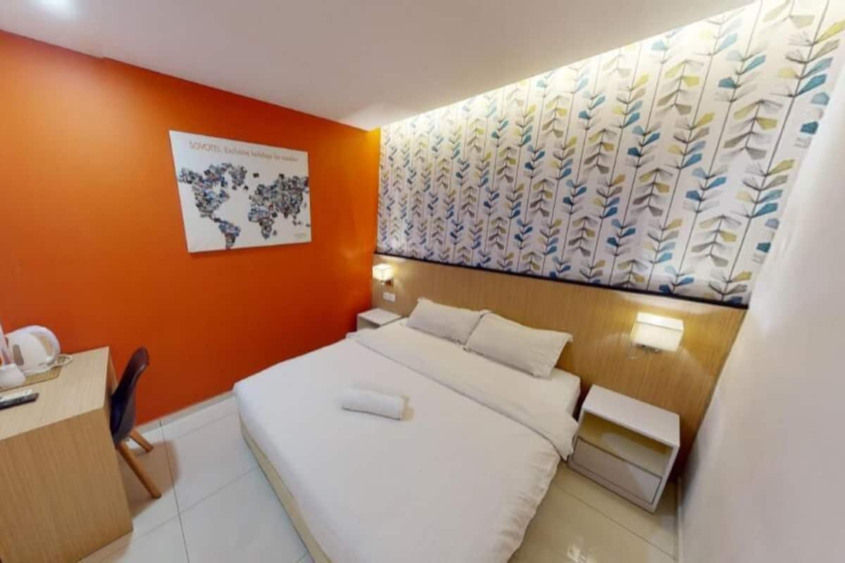 Ein Hotelzimmer mit einer orangefarbenen Akzentwand, einem Bett mit weißen Leinen, einem Schreibtisch mit Stuhl, einem Nachttisch und einem an der Wand befestigten Fernseher.