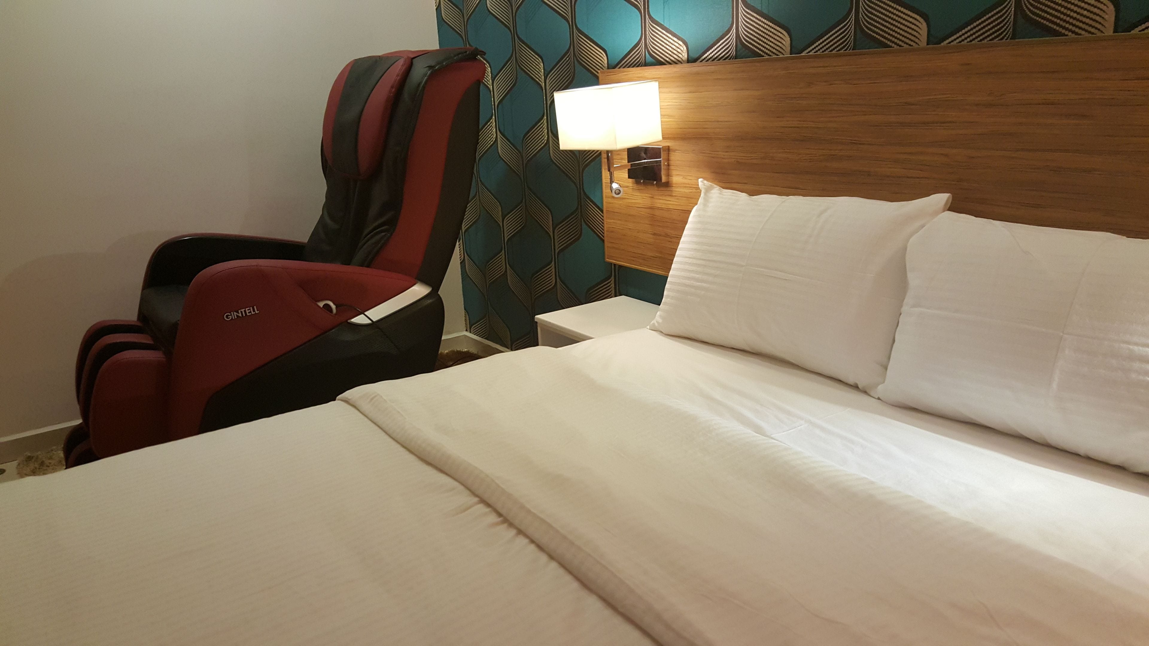A hotel room with a bed, a nightstand with a lamp, a red and black chair, and a patterned wall.