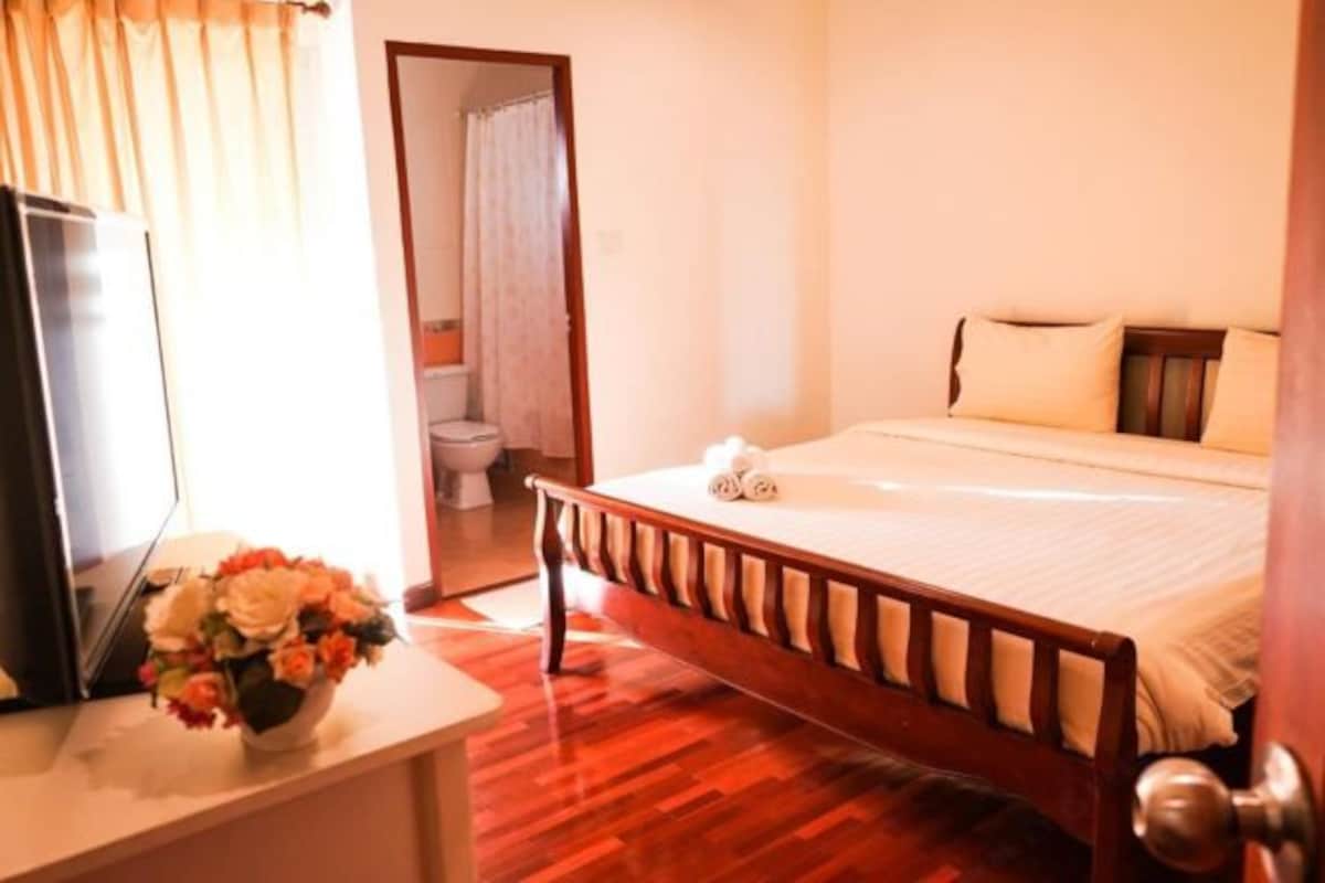 A hotel room with a bed, a bedside table with a vase of flowers, a television, and a wooden floor.