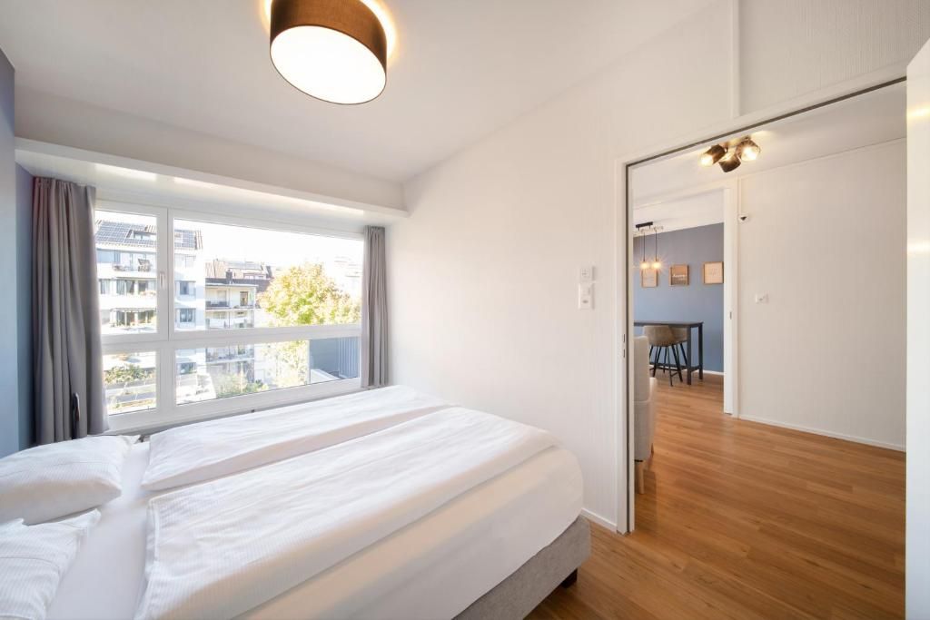 A bedroom with a large bed, a window with curtains, a wooden floor, and a door leading to a dining area.