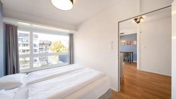 A bedroom with a large bed, a window with curtains, a wooden floor, and a door leading to a dining area.