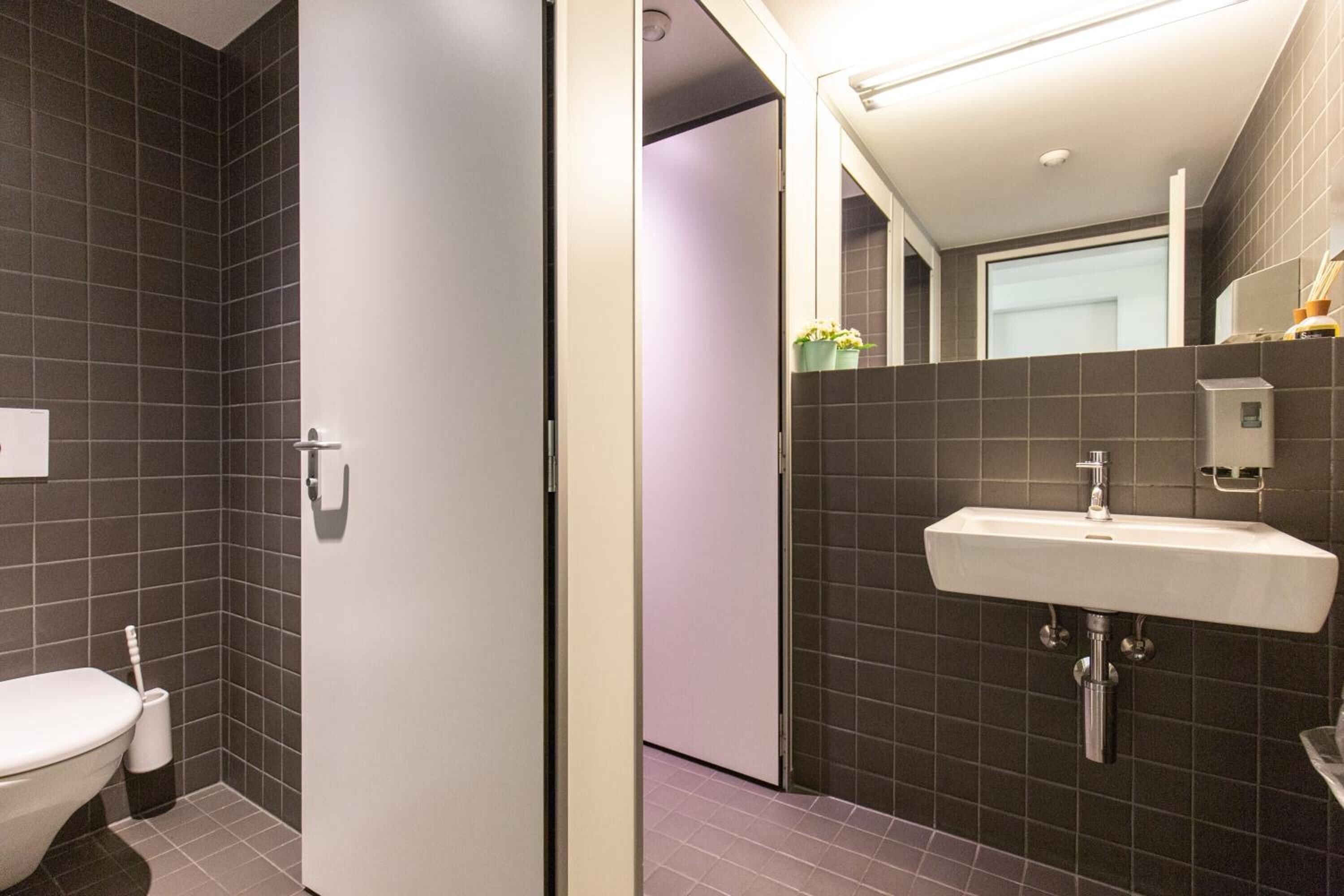 Ein modernes Badezimmer mit dunkel gefliesten Wänden, einer weißen Toilette und einem integrierten Waschbecken mit Spiegel darüber.