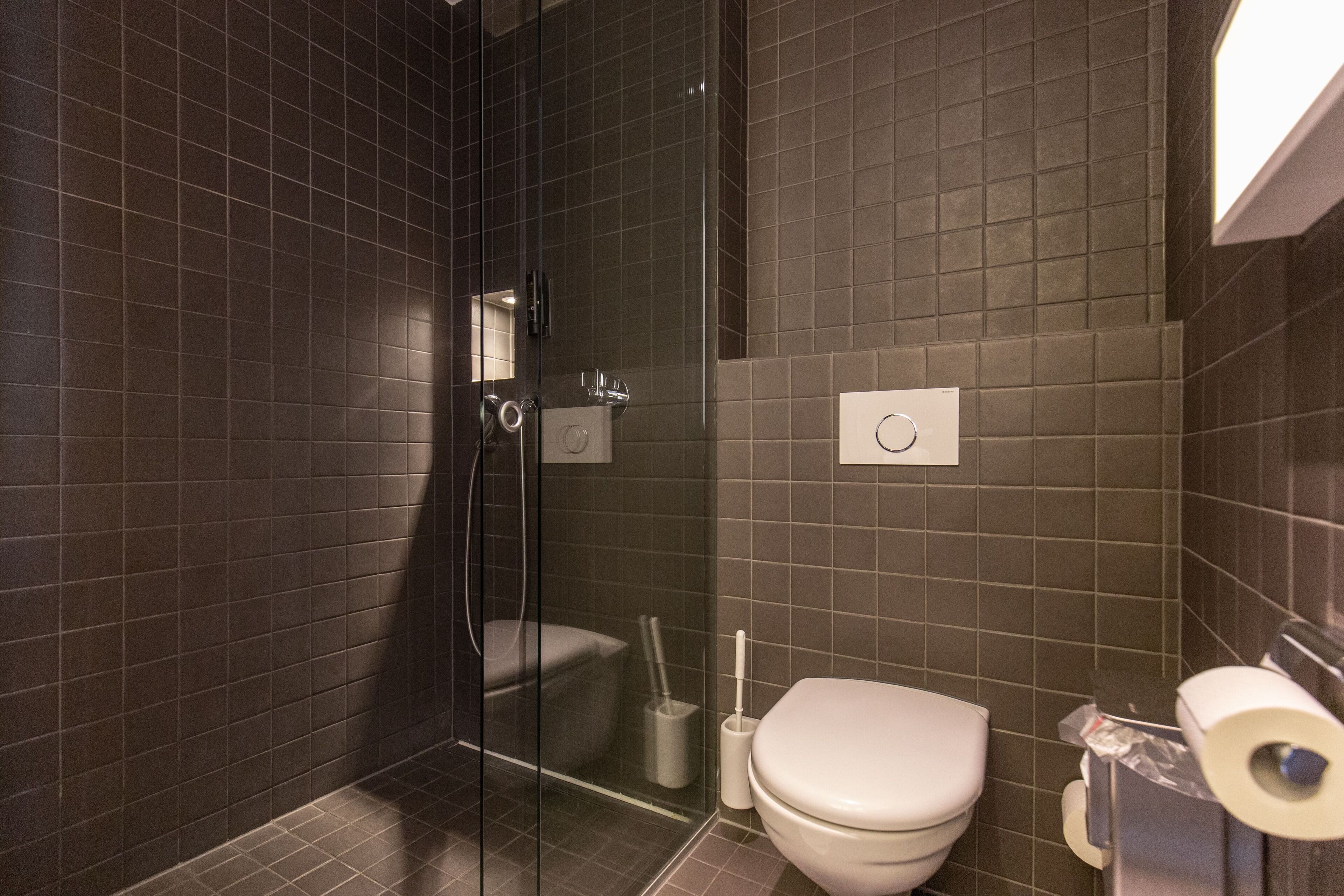 Un baño moderno con baldosas oscuras, cabina de ducha de vidrio, inodoro y dispensador de toallas de papel.
