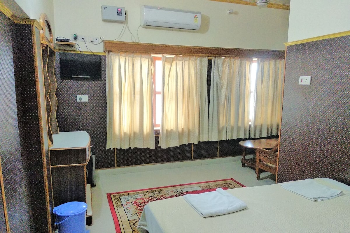 A hotel room with a bed, a desk, a chair, a television, and an air conditioning unit.