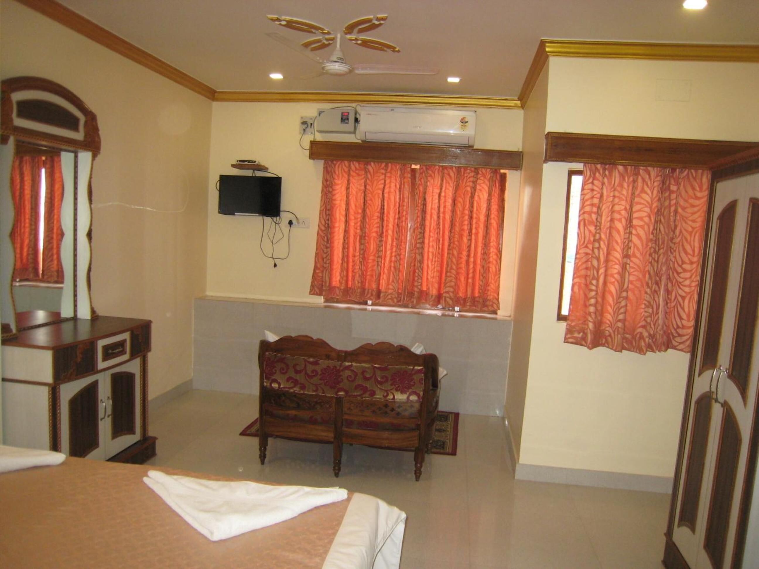 A hotel room with a bed, a wooden dresser, a TV mounted on the wall, a window with orange curtains, and a ceiling fan.