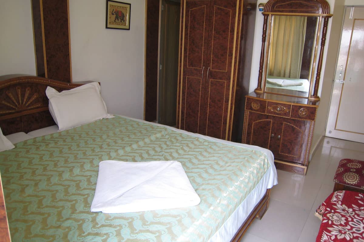 A neatly made bed with a patterned bedspread, a wooden wardrobe, a mirror with a curtain, and a red patterned rug.
