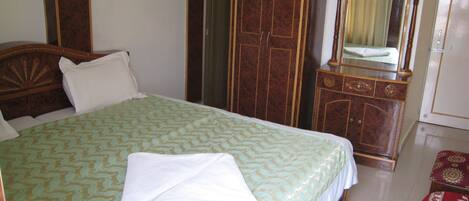 A neatly made bed with a patterned bedspread, a wooden wardrobe, a mirror with a curtain, and a red patterned rug.