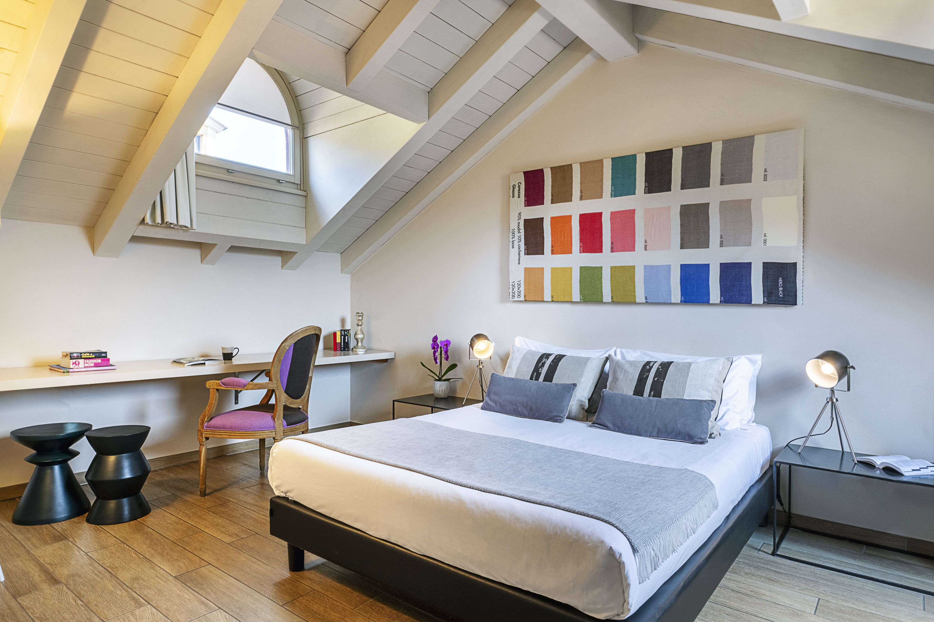 Een moderne slaapkamer met een loft-plafond, een groot bed met veel kussens, een bureau met stoel en een kleurenpalet op de muur.