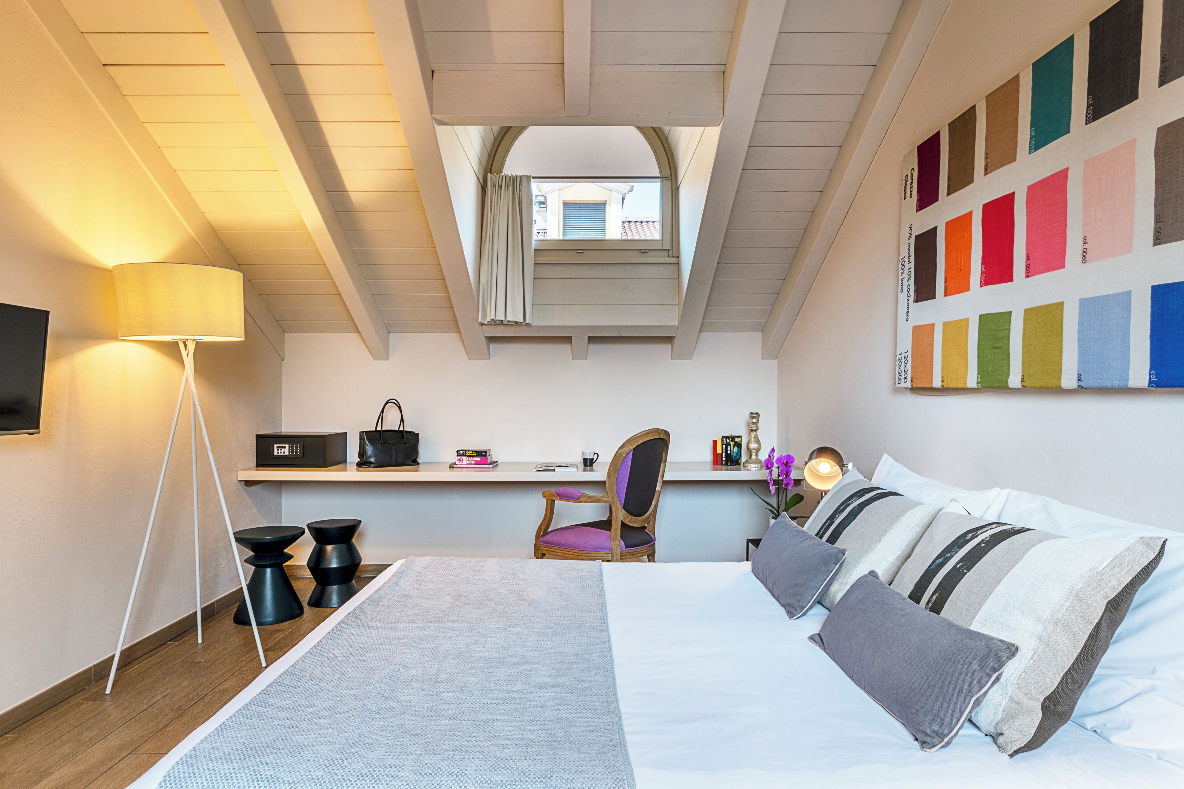 Un dormitorio moderno con tragaluces, una cama con ropa de cama gris y blanca, una mesita de noche con lámpara y una paleta de colores en la pared.