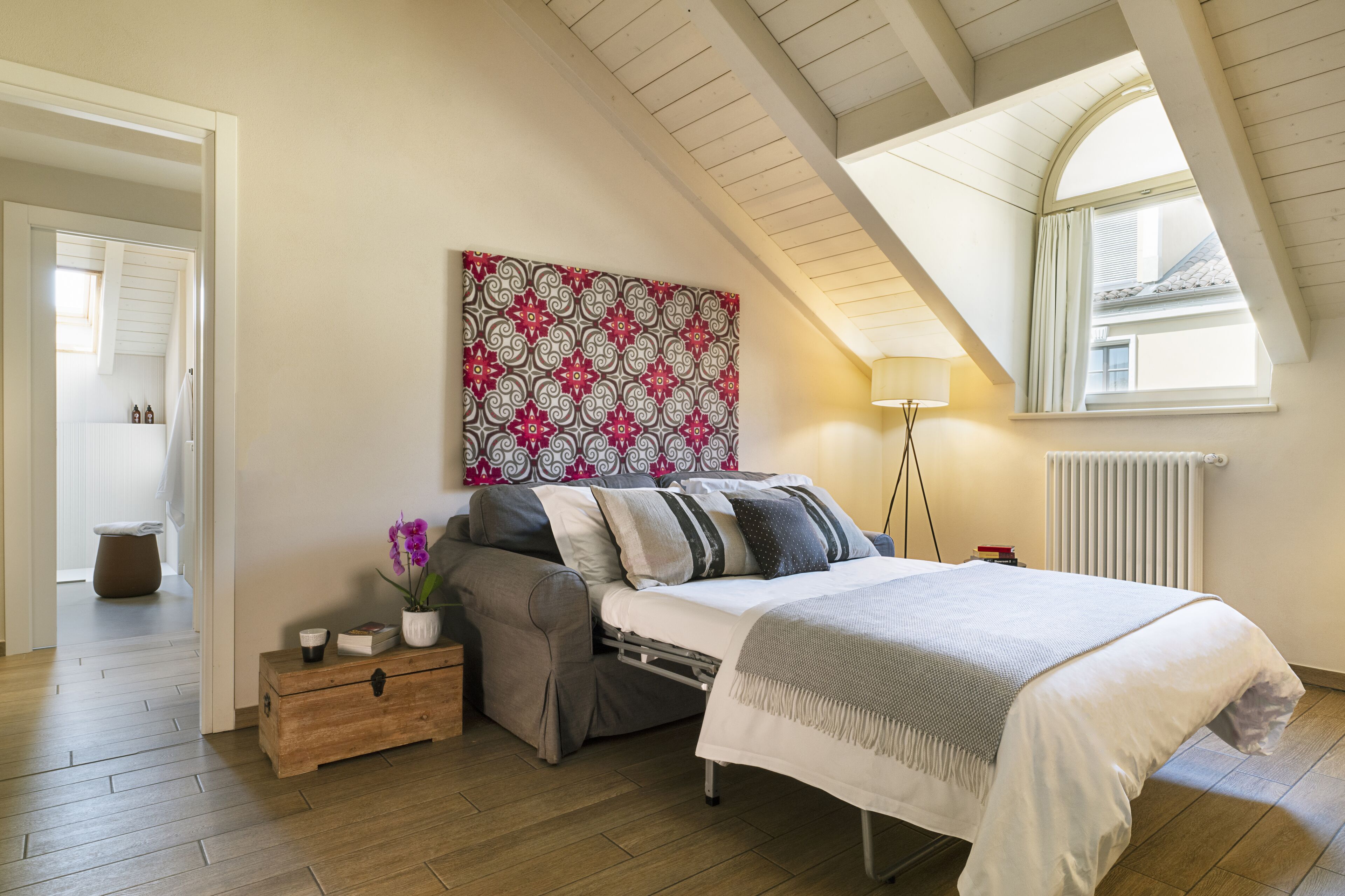 A bedroom with a loft-style ceiling, a bed with a white comforter, a bedside table with a lamp, a decorative wall piece, and a view of the outside through a window.