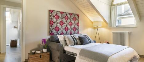 A bedroom with a loft-style ceiling, a bed with a white comforter, a bedside table with a lamp, a decorative wall piece, and a view of the outside through a window.