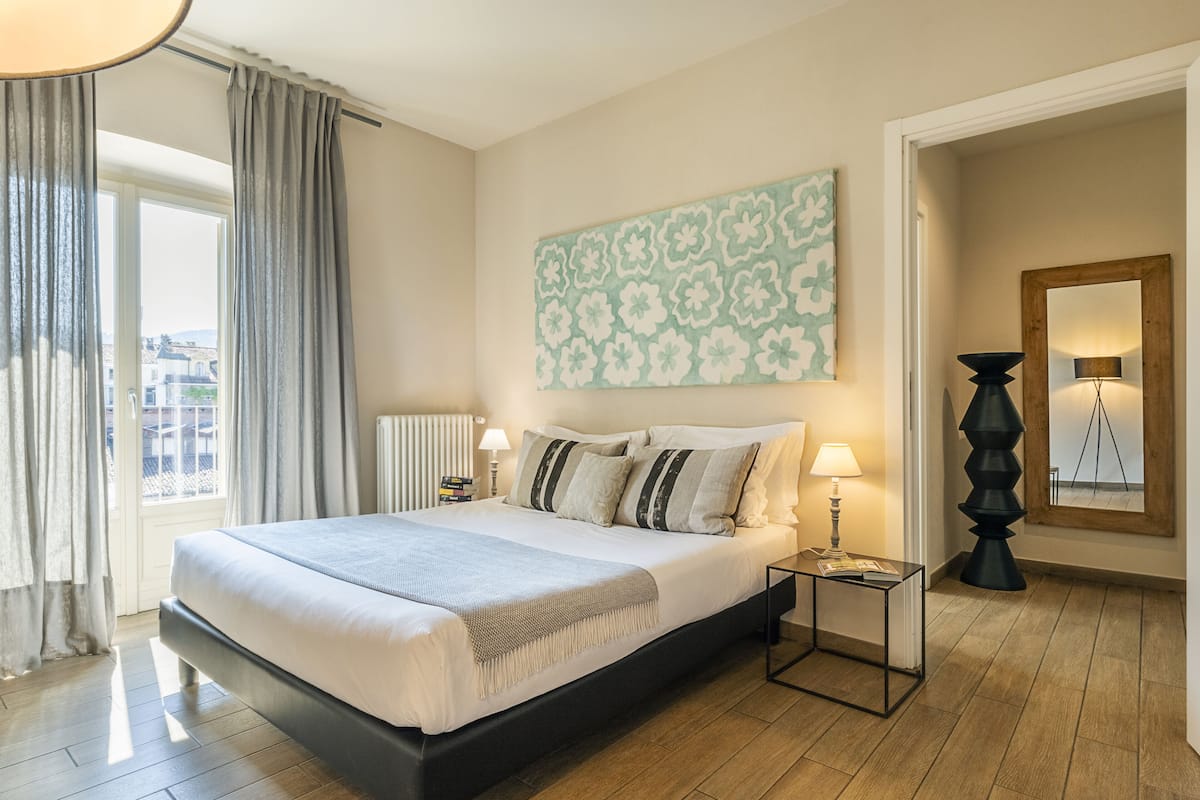 Un dormitorio con una cama grande, dos mesitas de noche con lámparas, una ventana con cortinas, una obra de arte colgada en la pared y un espejo con marco de madera.