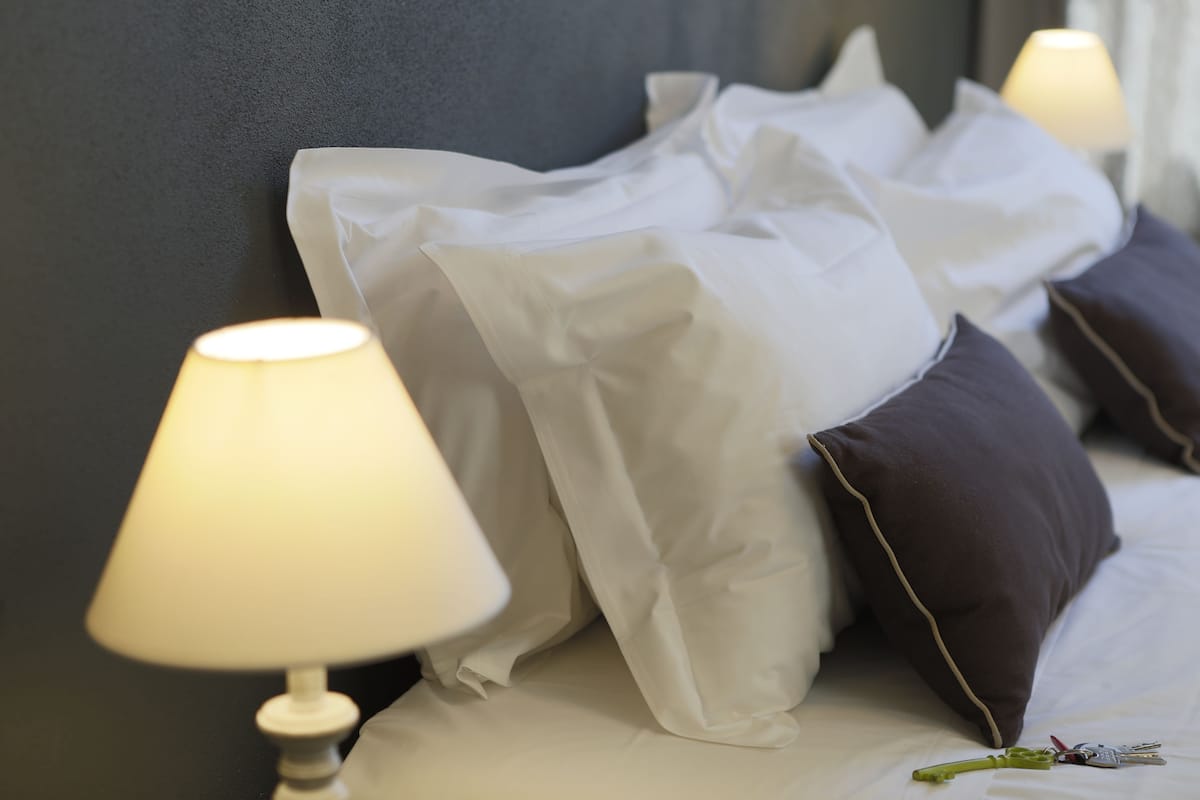 Una cama bien hecha con almohadas blancas y oscuras, una lámpara a un lado y un llavero sobre la cama.