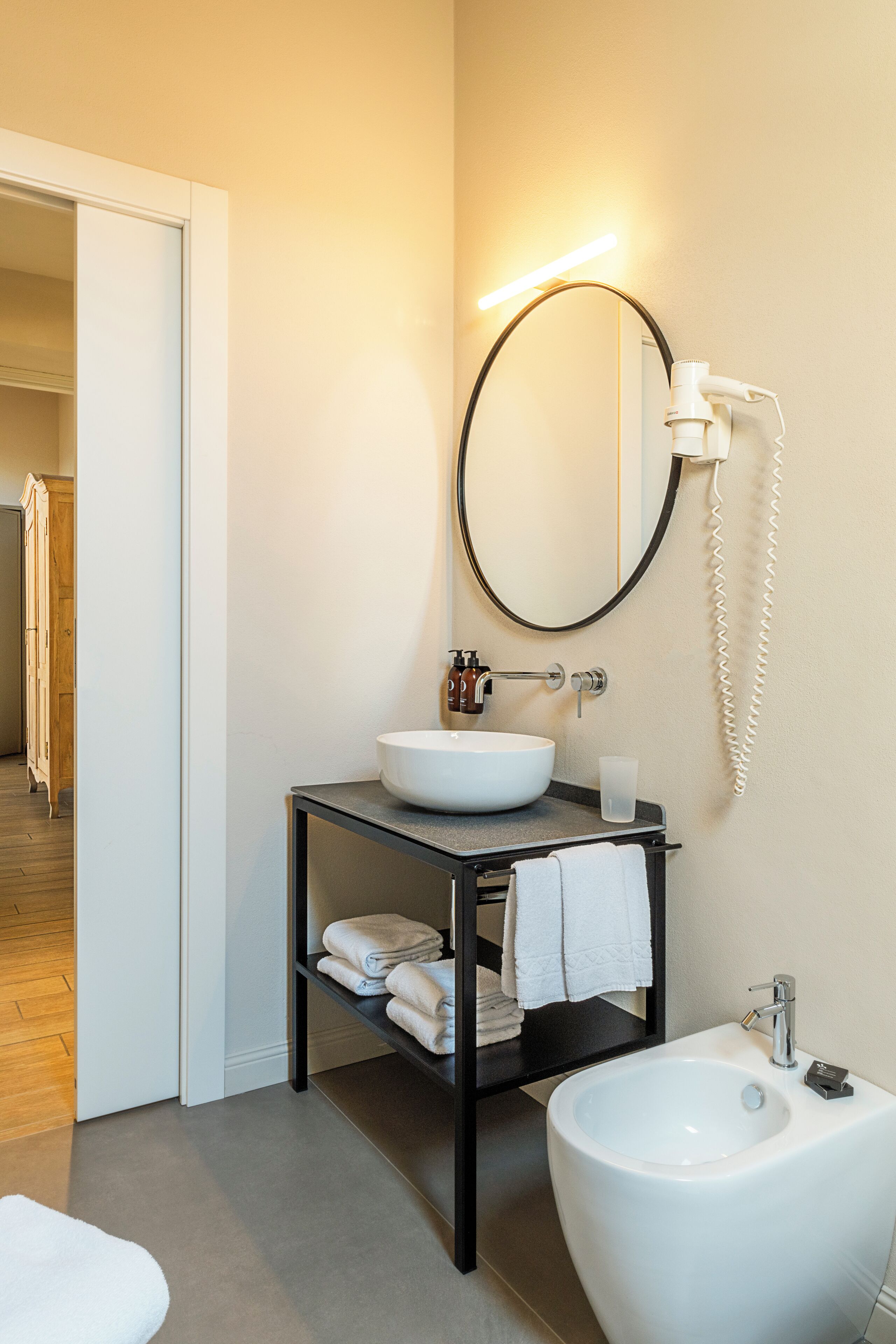 Un bagno moderno con un lavandino bianco, un bidet e uno specchio. È presente un portasciugamani con asciugamani piegati e un piccolo ripiano con articoli da toilette.