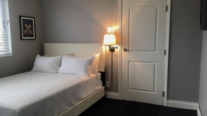 A neatly made bed with white linens in a room with grey walls, a bedside lamp, and a door. - Ivoire Guest House (Sekondi-Takoradi)