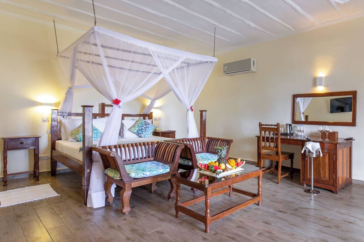 Una habitación con una cama, dos sillas de madera, una mesa con fruta, un espejo y una mesita de noche con una lámpara.
