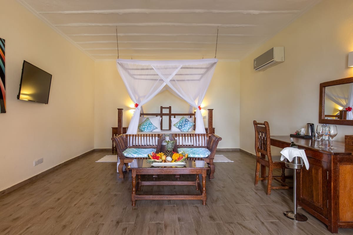 Una habitación con un banco de madera, una pequeña mesa con fruta, una cama con dosel, una silla de madera, una zona de bar y un televisor montado en la pared.