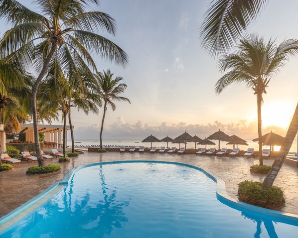 Ein Poolbereich mit Liegestühlen und Sonnenschirmen, umgeben von Palmen und Blick auf das Meer bei Sonnenuntergang.