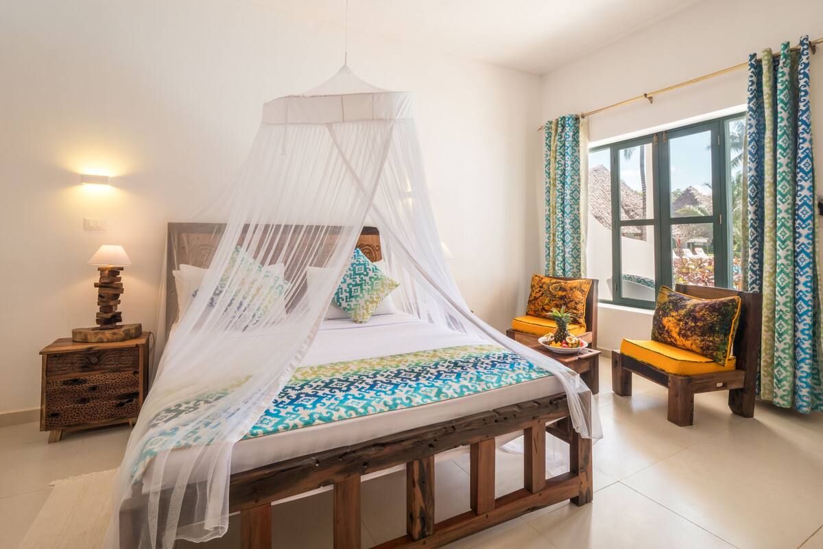 Un dormitorio con cama con dosel, una mesita de noche con lámpara, una silla con cojín y una ventana con cortinas.