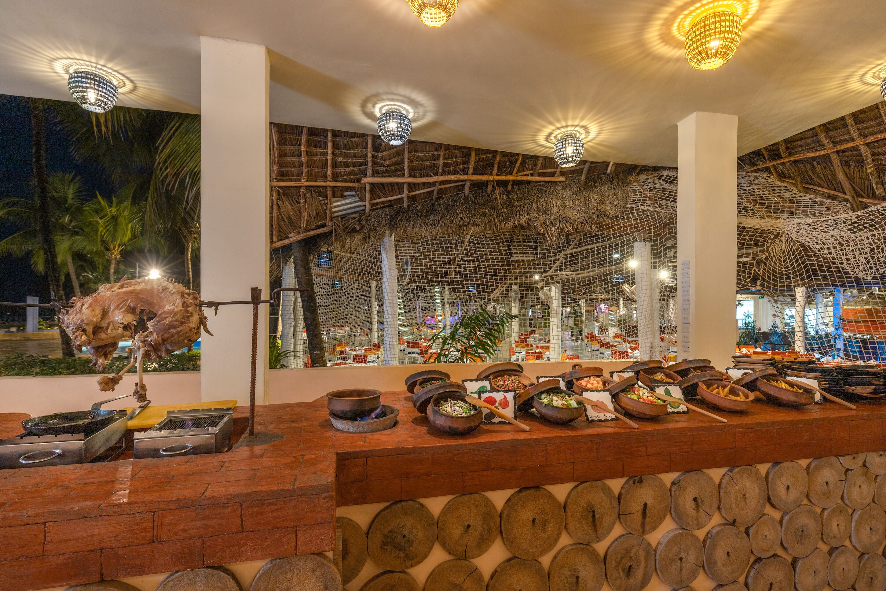 A restaurant with a thatched roof, a variety of dishes displayed on a counter, and a hanging meat preparation.