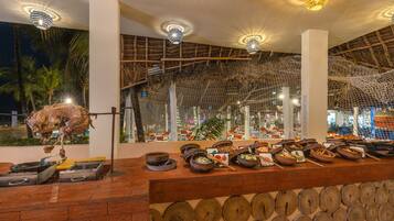 A restaurant with a thatched roof, a variety of dishes displayed on a counter, and a hanging meat preparation.