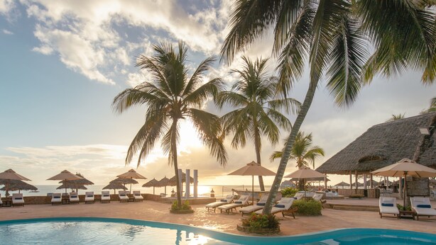 Un área de piscina con sillones de descanso y sombrillas, palmeras y vistas al atardecer.