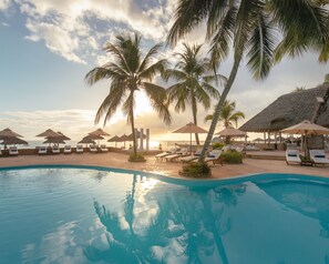 Ein Poolbereich mit Liegestühlen und Sonnenschirmen, Palmen und Blick auf den Sonnenuntergang.