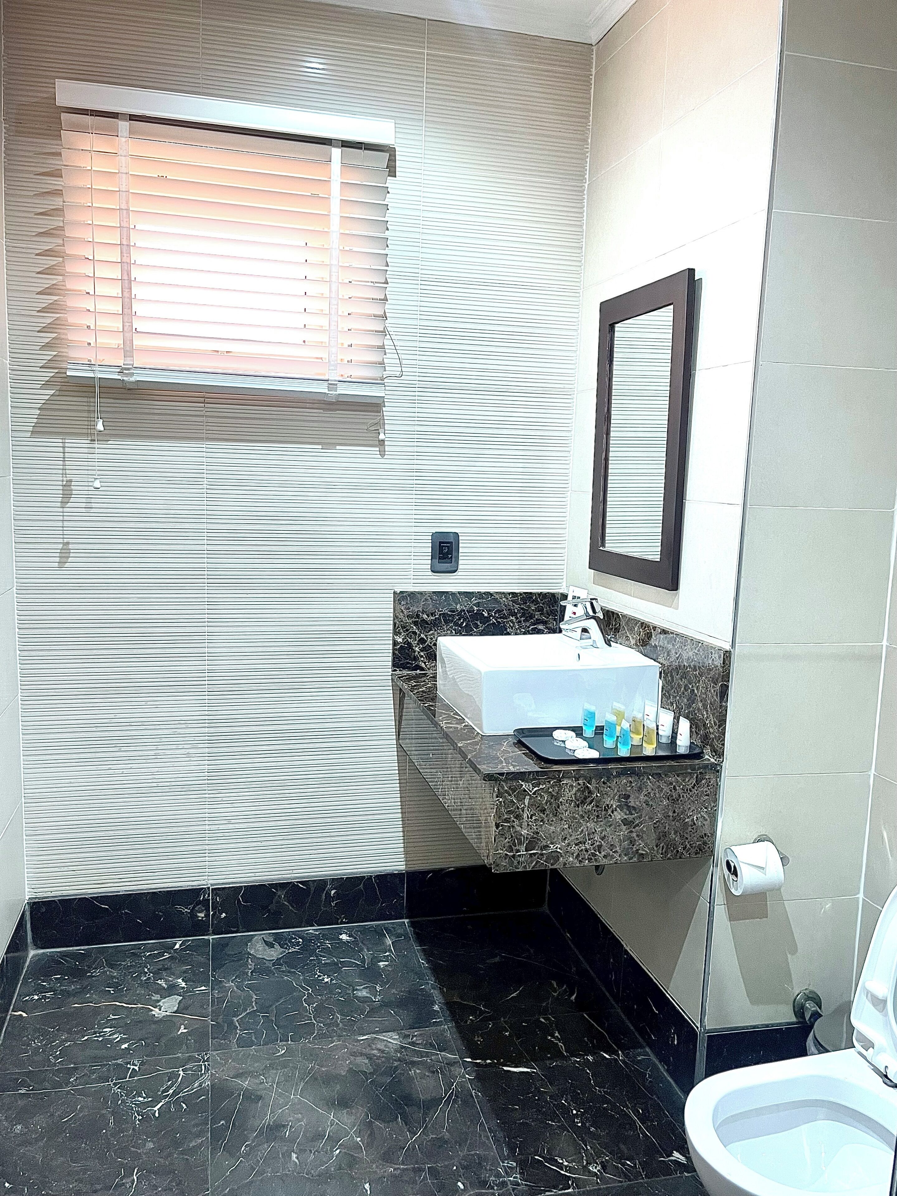 Un baño con suelo de mármol, un lavabo blanco, un espejo y una ventana con persianas.