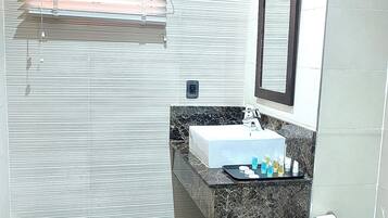 Un baño con suelo de mármol, un lavabo blanco, un espejo y una ventana con persianas.