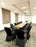 Un salón de conferencias con una larga mesa, sillas negras, una pantalla de proyector y persianas en las ventanas.