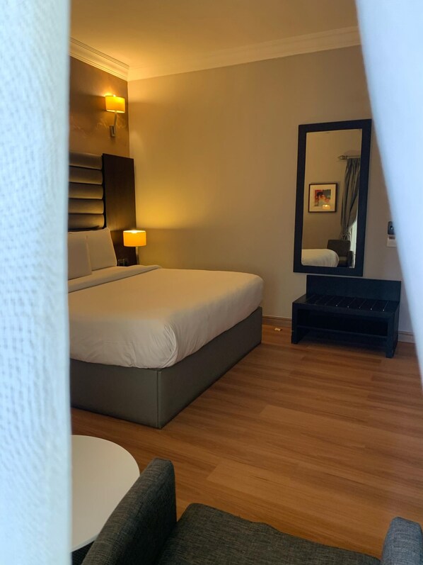 A modern hotel room with a large bed, bedside lamps, a small table, a chair, a mirror, and a wooden floor.