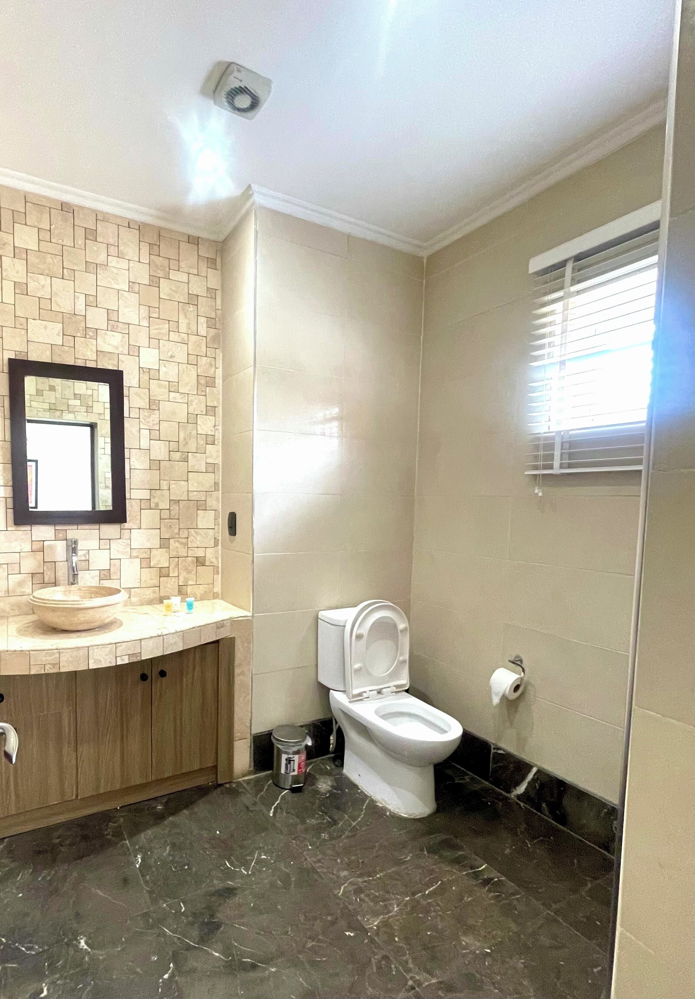 A bathroom with a beige tiled wall, a sink with a mirror, a toilet, and a window with blinds.